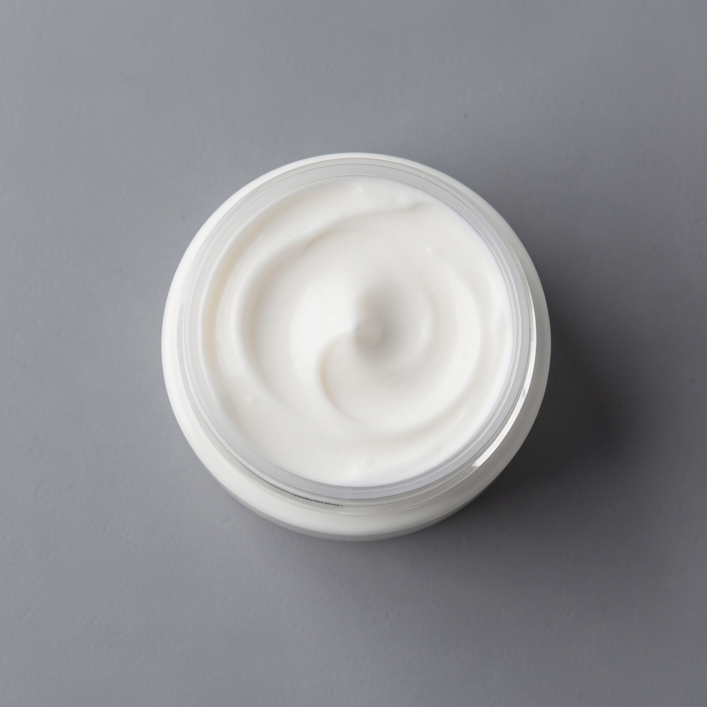 Open round jar of thick rich body butter in warm cream tone, flat lay on pale grey surface with botanical leaf accent